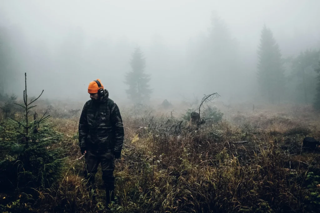 A person wearing a bright orange beanie, headphones, and a dark jacket stands in a foggy, overgrown field looking down. The background features tall, hazy evergreen trees shrouded in dense fog.
