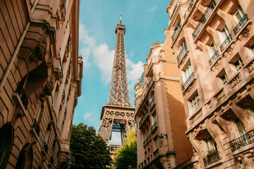 A low-angle view looking up a narrow Parisian street framing the Eiffel Tower against a bright, clear blue sky. Warmly lit classic Haussmann-style residential buildings line both sides of the street with detailed balconies and stone facades.