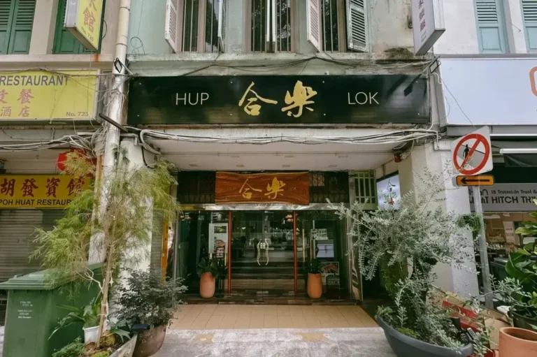 A traditional storefront features prominent black and wooden signage reading "HUP LOK" alongside Chinese characters above glass entrance doors. Potted plants flank the entrance, set against the street-level facade of a multi-story building.