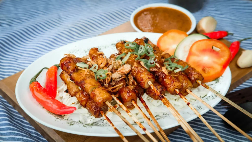 A white plate holds grilled skewers topped with fried shallots and green onions, served alongside fresh tomato and cucumber slices. In the background, red chili peppers and a small bowl of savory dipping sauce rest on a wooden board and blue striped tablecloth.