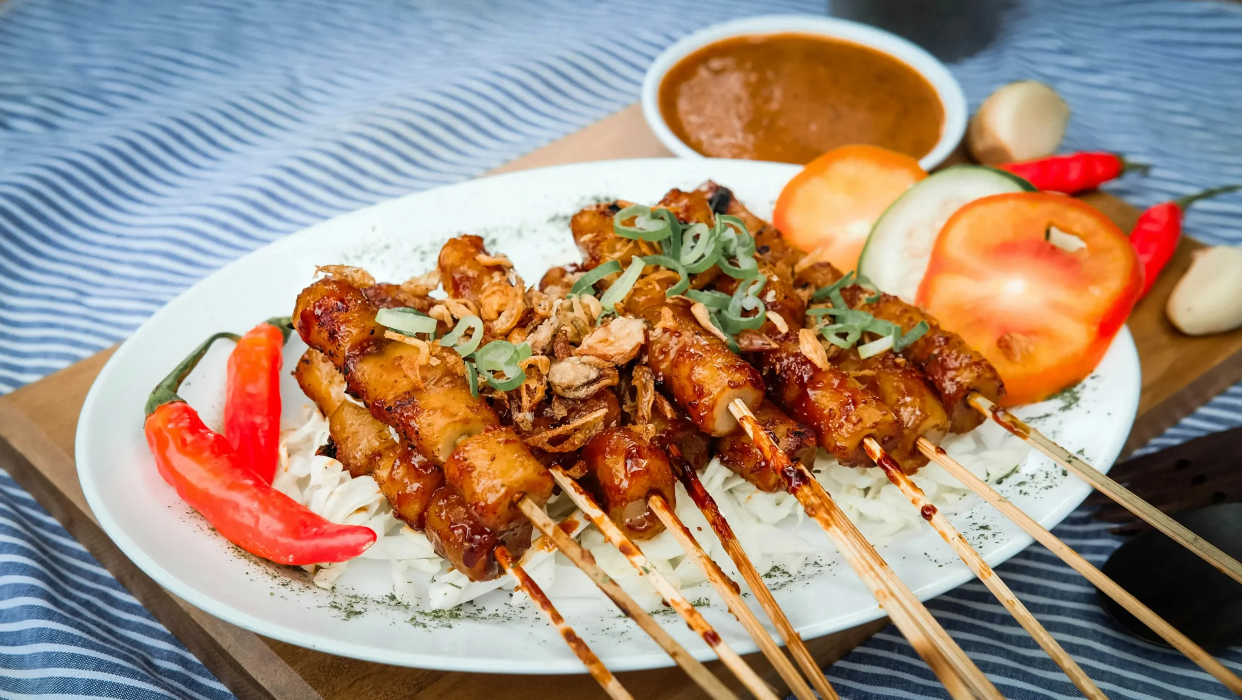 A white plate holds grilled skewers topped with fried shallots and green onions, served alongside fresh tomato and cucumber slices. In the background, red chili peppers and a small bowl of savory dipping sauce rest on a wooden board and blue striped tablecloth.