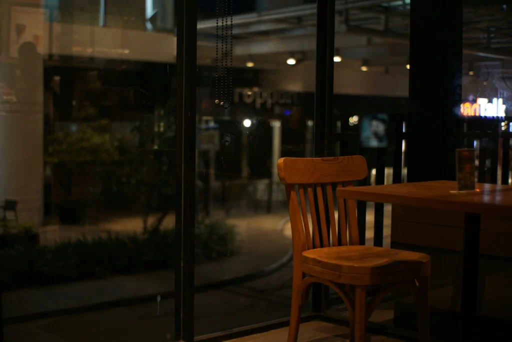 A wooden chair sits next to a table inside a dimly lit cafe at night, positioned right in front of a large window. Through the glass, glowing city streetlights and store signs create a moody backdrop.