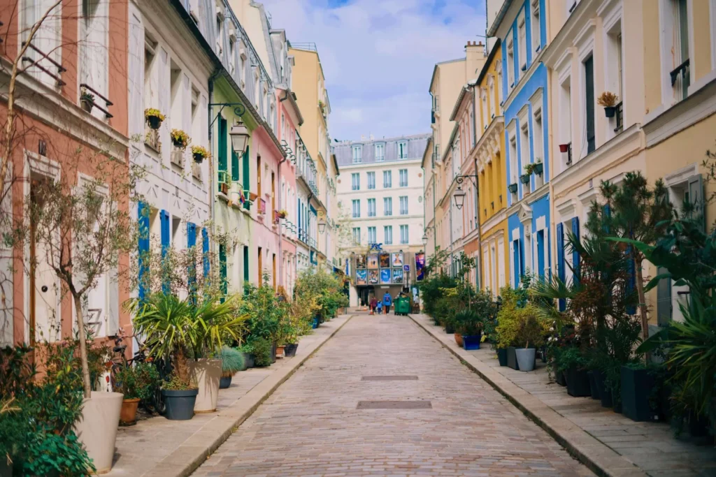 A cobblestone residential street is lined with colorful, pastel-painted buildings and lush potted plants on both sides. The narrow pathway stretches toward a white building at the end under a partly cloudy blue sky.