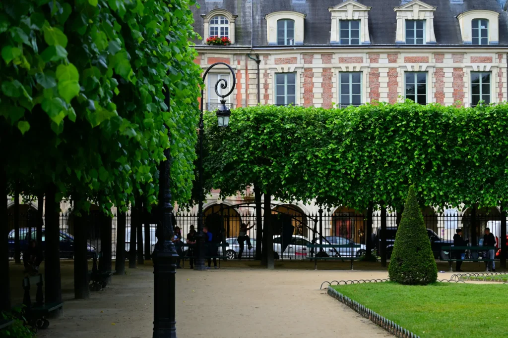 A tree-lined walkway with neatly manicured foliage and a black ornate lamppost stands in the foreground of a park. In the background, elegant brick and stone European-style buildings feature large rectangular and dormer windows.