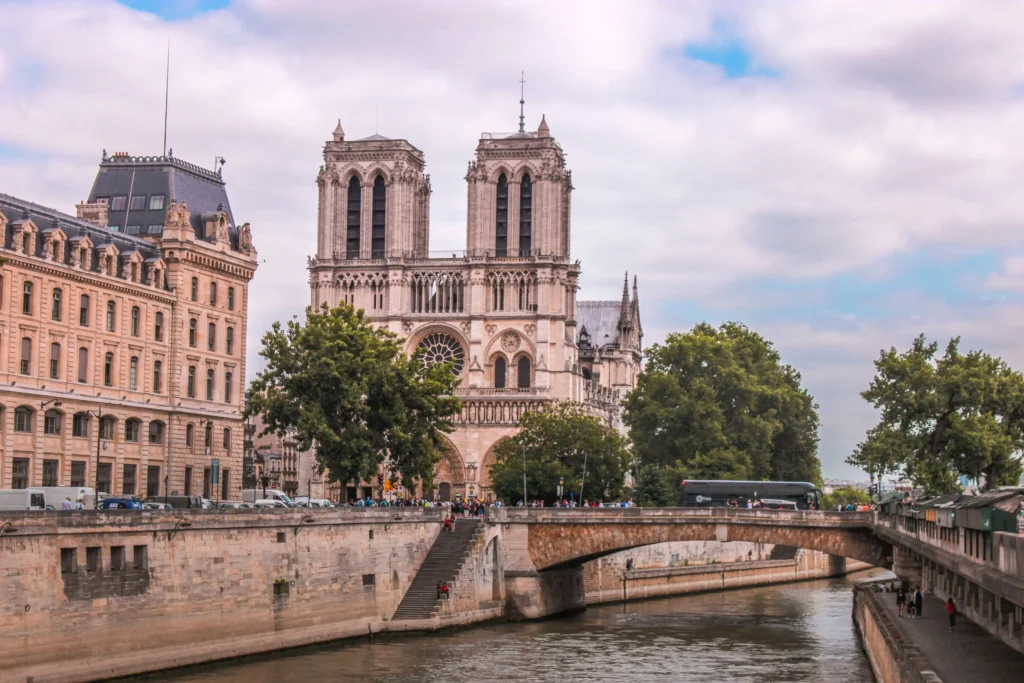A scenic daytime view of the iconic Notre-Dame Cathedral in Paris standing along the edge of the Seine River. An arched stone bridge crosses the calm water in the foreground under a lightly clouded sky.