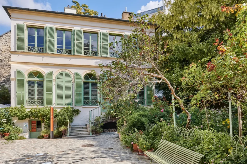 A two-story white building with sage green shutters overlooks a stone courtyard surrounded by lush greenery and leafy trees. A matching green wooden bench sits in the garden foreground near a small staircase leading up to the building's entrance.