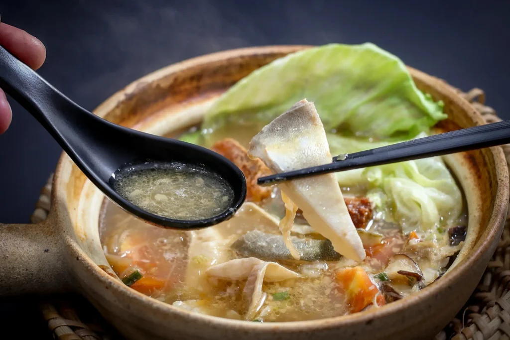A clay pot filled with hot pomfret fish soup containing green cabbage leaves, mushrooms, and tomatoes. Black chopsticks lift a piece of white fish from the broth while a black spoon holds a spoonful of the warm soup.