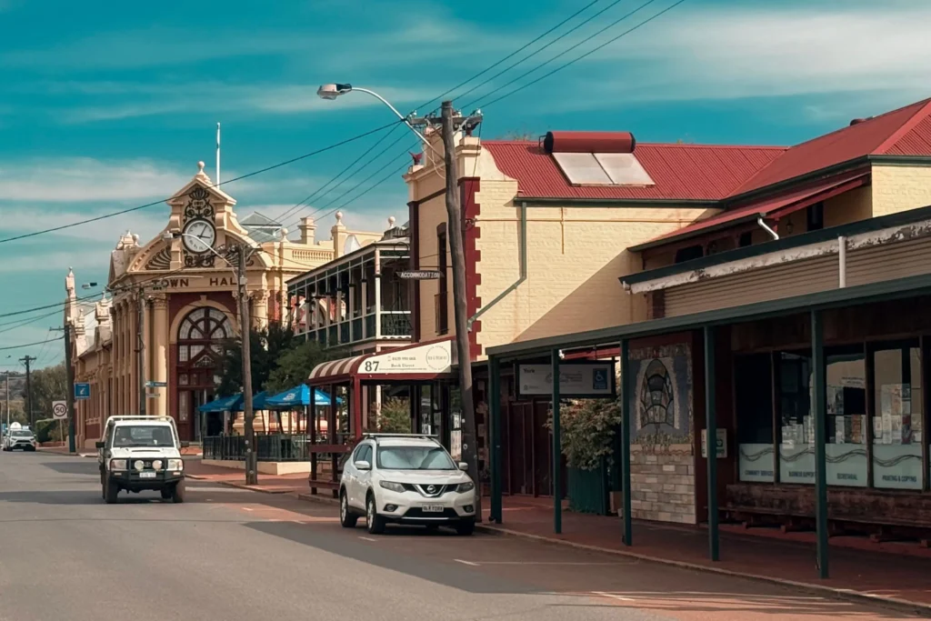 A sunlit street scene in a historic town featuring classic brick and stone buildings with red tin roofs and covered storefront walkways. A white SUV is parked along the curb, and a white off-road vehicle drives down the road near a prominent Town Hall clock tower.