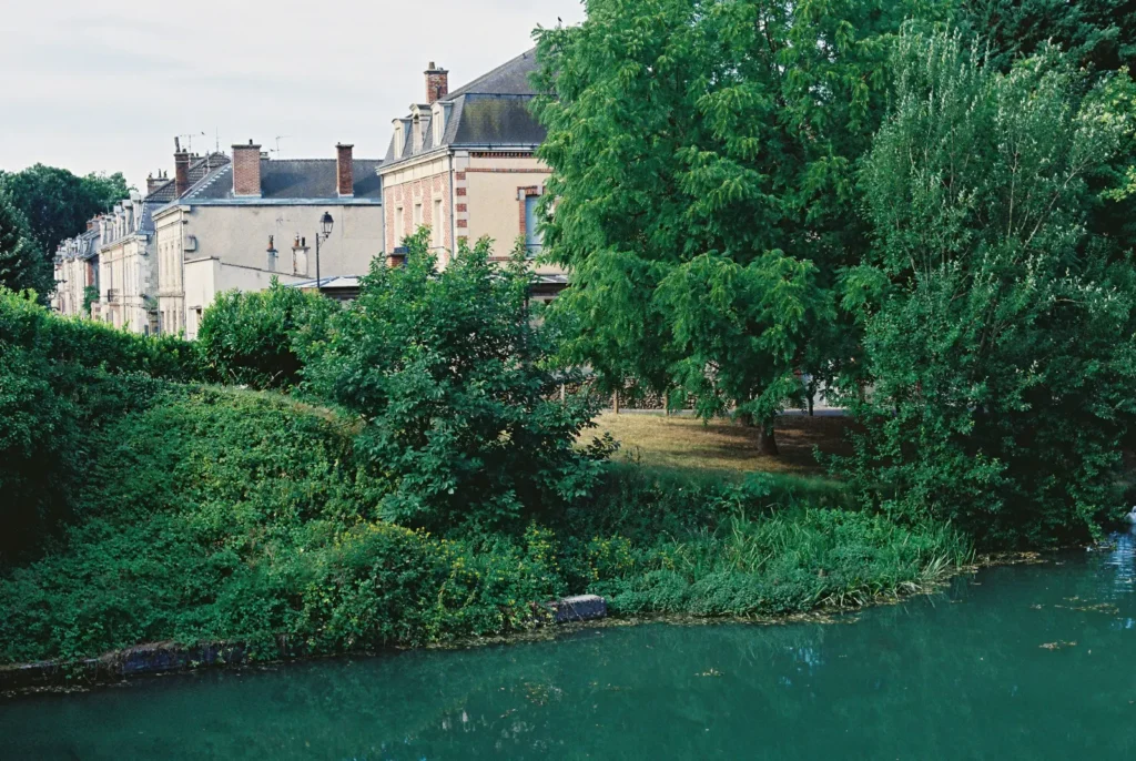 A row of European-style buildings with brick accents stands beside a calm, green canal. Lush leafy trees and dense shrubbery cover the riverbank in the foreground.