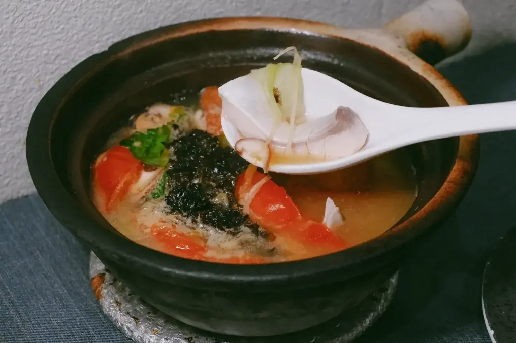 A white ceramic soup spoon scoops up a piece of tender fish, green scallion, and shredded ginger from a steaming clay pot dish. The dark earthenware pot is filled with a rich broth, sliced tomatoes, seaweed, and fresh cilantro.
