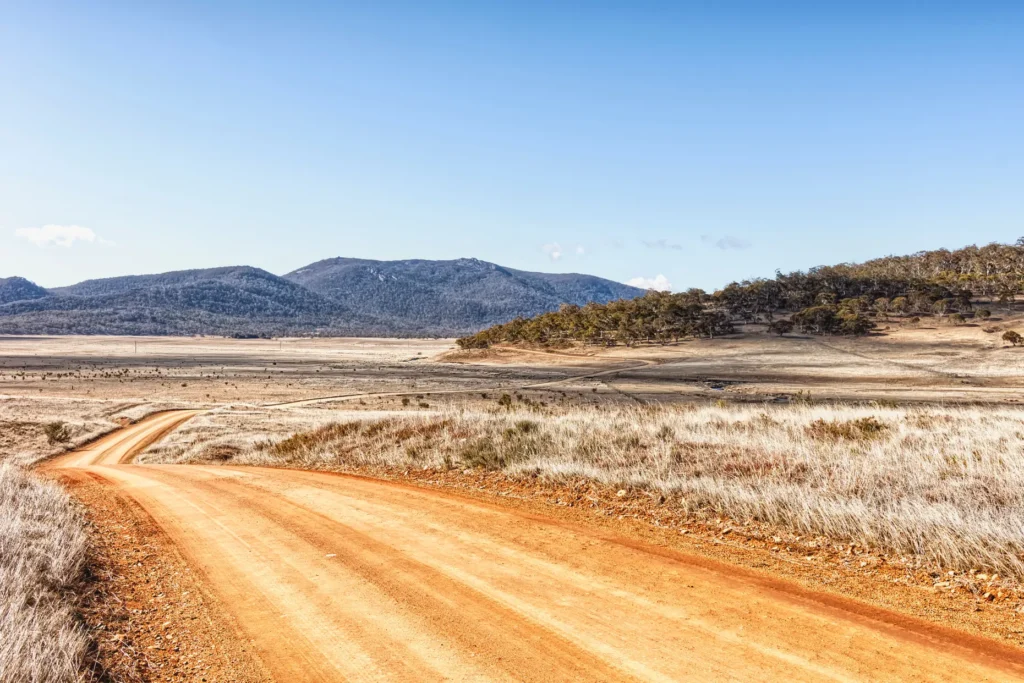 A winding orange dirt road stretches through a dry, grassy countryside under a clear blue sky. In the background, rolling hills and scattered trees frame the wide open landscape.