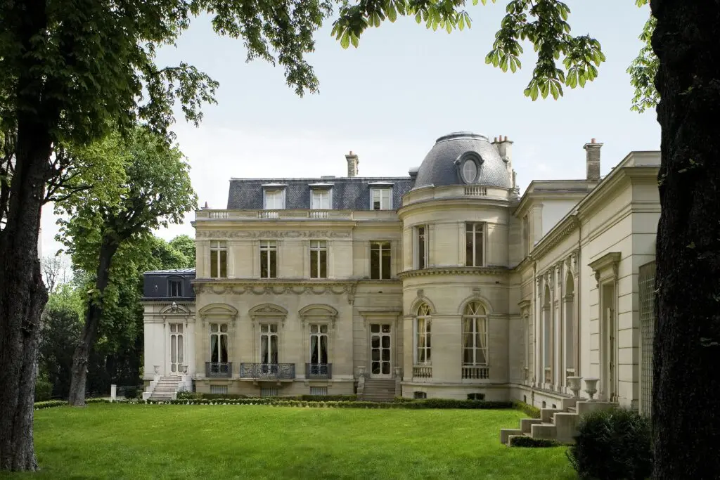 Exterior view of the Musée Marmottan Monet, a grand French mansion featuring light beige stone facades and a domed tower section. Lush green grass and leafy mature trees frame the elegant grounds surrounding the historic building.