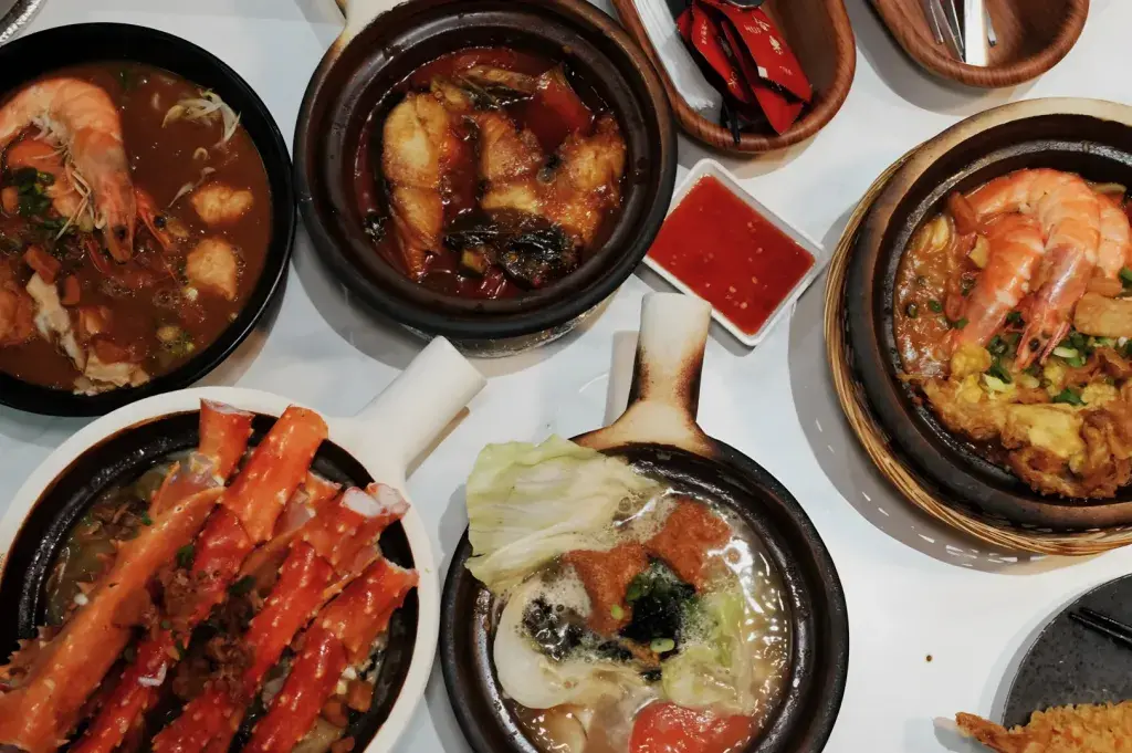 An overhead view of a table filled with various clay pot dishes containing seafood, including crab legs, whole shrimp, and fish fillets in rich sauces. The vibrant spread features clear broths, spicy red gravies, and a side of red dipping sauce arranged neatly on a white surface.