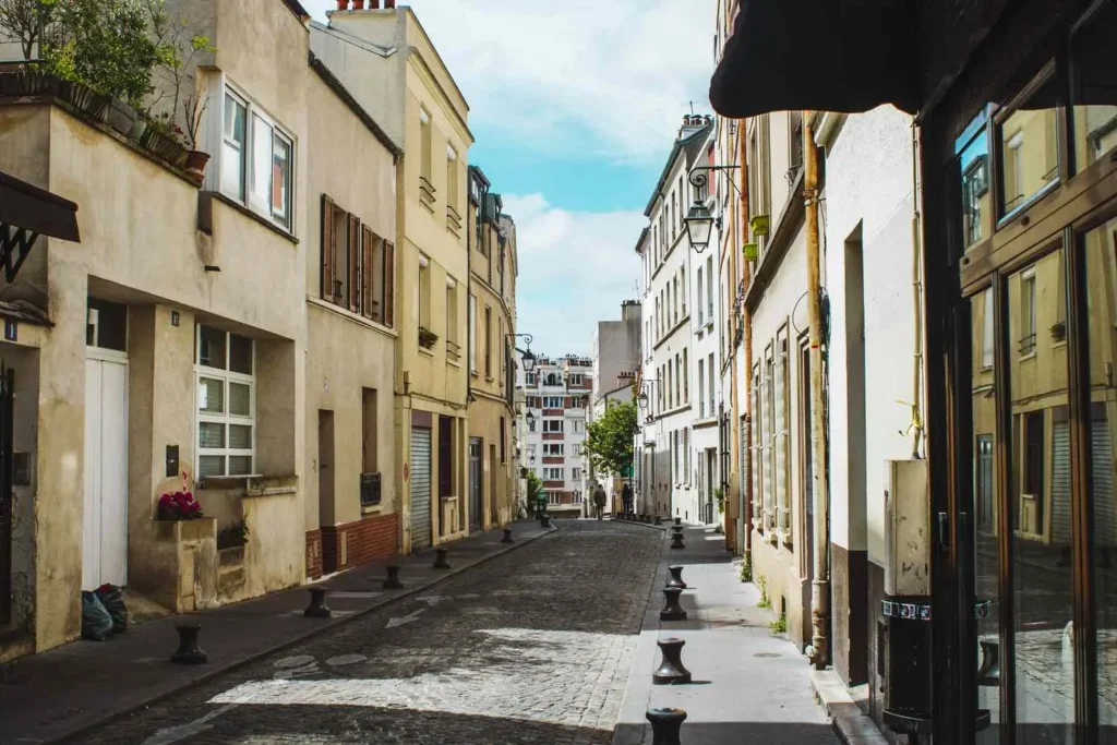A narrow, cobblestone European street winds between multi-story residential buildings bathed in warm sunlight. Small bollards line the narrow sidewalks under a partly cloudy sky.