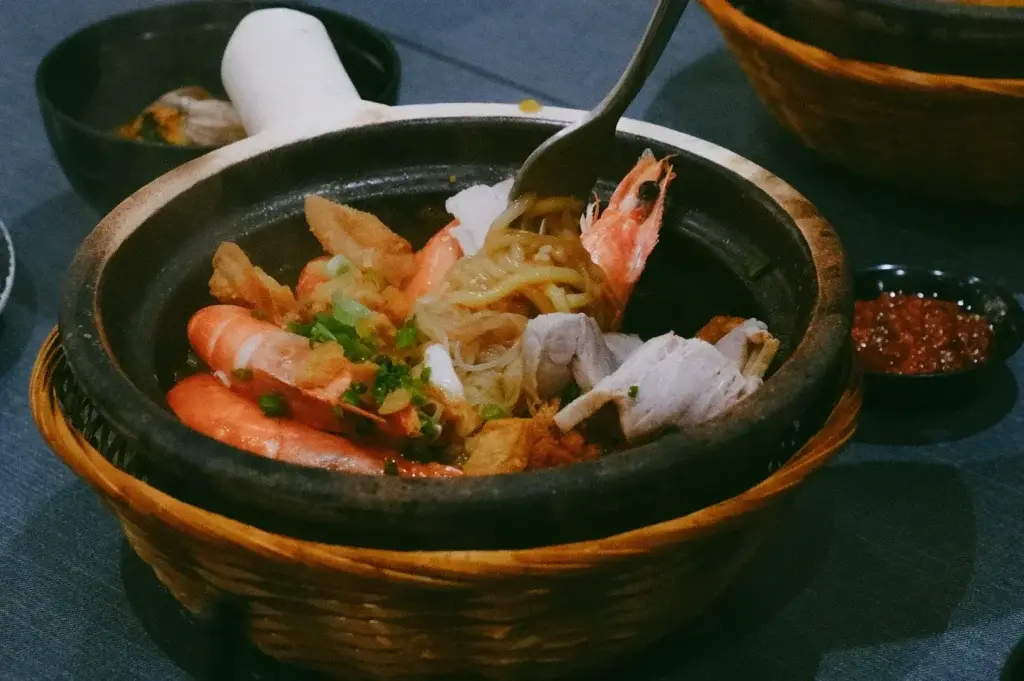 A hot clay pot filled with noodles and fresh seafood sits in a woven basket standard. A metal spoon lifts a portion of noodles surrounded by whole whole-head prawns, seafood chunks, chopped scallions, and fried dough pieces.