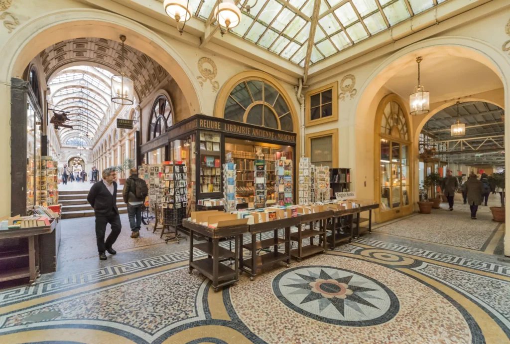 A sunlit, historic shopping arcade with vaulted glass ceilings features a mosaic-tile floor with a prominent compass star pattern. People stroll past classical storefronts displaying rows of books and postcards outside a vintage bookstore.