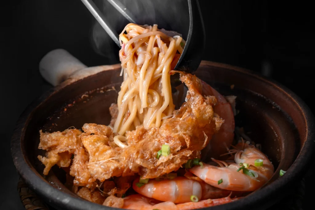 A clay pot filled with Hokkien mee noodles, cooked prawns, and crisp crispy lard or fried egg topping against a dark background. Black tongs lift a steaming clump of noodles directly from the dish.