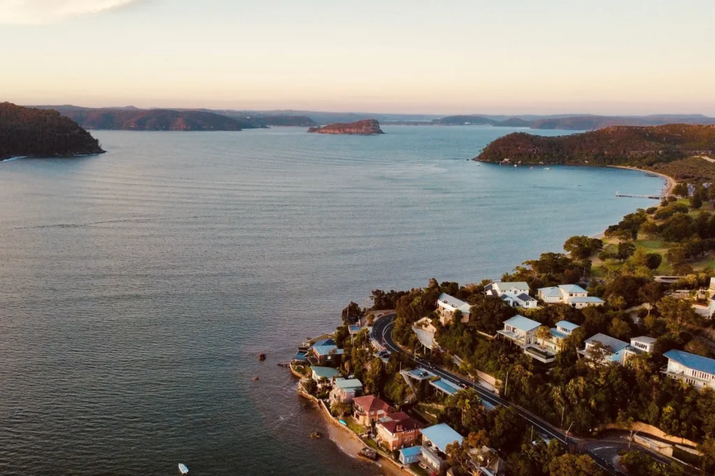 An aerial view captures a coastal town nestled among lush green hills bordering a wide, calm bay. Residential houses and a winding road line the shoreline under the warm glow of a setting sun.