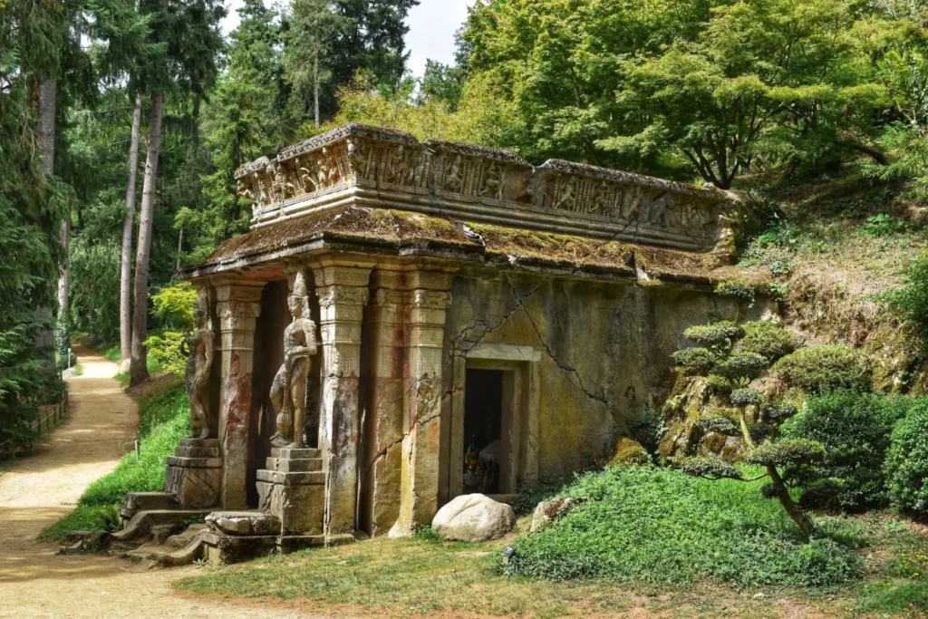 An ornate stone structure featuring detailed carvings, decorative columns, and carved figures stands built into a mossy, wooded hillside. A dirt path winds alongside the lush greenery and surrounding trees next to the ancient-style monument.