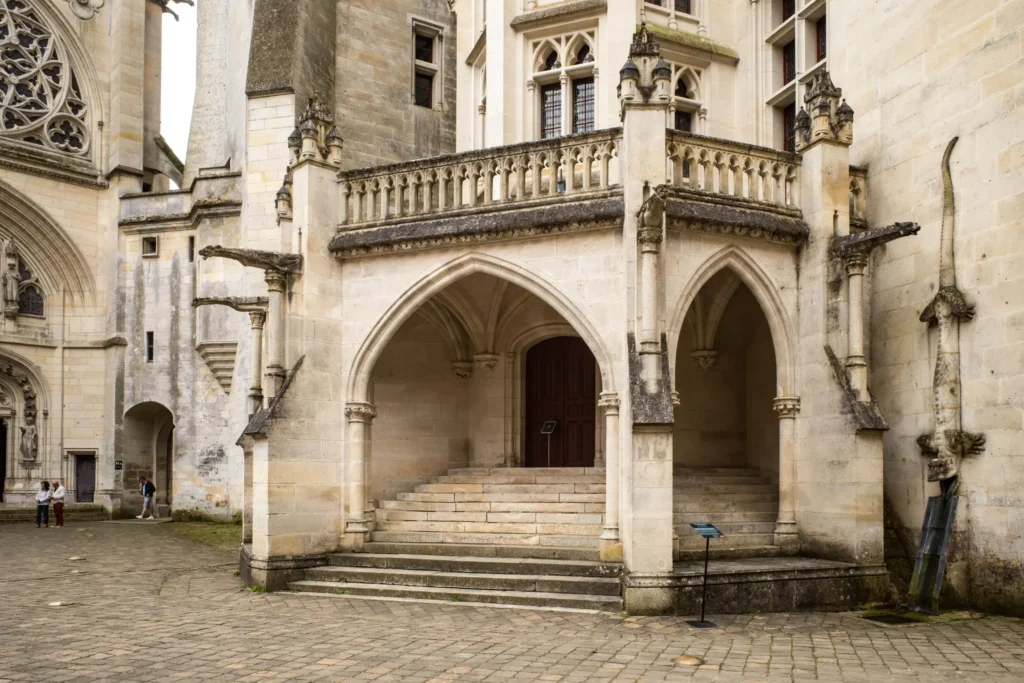 A view of a courtyard featuring a stone architectural entrance with pointed arches, steps, and decorative gargoyles. On the far right wall, a detailed stone sculpture of a lizard-like creature climbs vertically down toward a gutter spout.