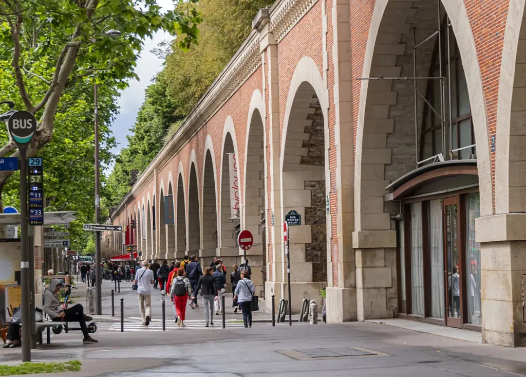 A side-angle view of the Viaduc des Arts in Paris showing a row of tall brick archways set along a tree-lined sidewalk. Pedestrians stroll along the walkway near a bus stop and street signs as greenery grows atop the structure.