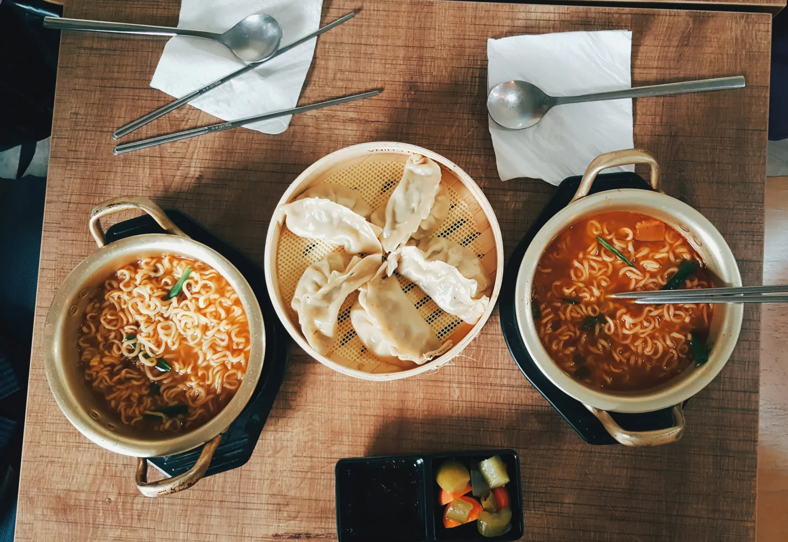 A top-down view of a wooden table set with two small brass pots of spicy ramen soup and a central steamer basket filled with dumplings. Metal chopsticks, spoons on white napkins, and a small side dish of pickled vegetables are neatly arranged around the food.