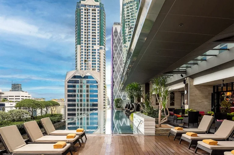 Rooftop swimming pool at Eastin Grand Hotel Sathorn with lounge chairs and yellow towels, surrounded by greenery and tall city buildings.