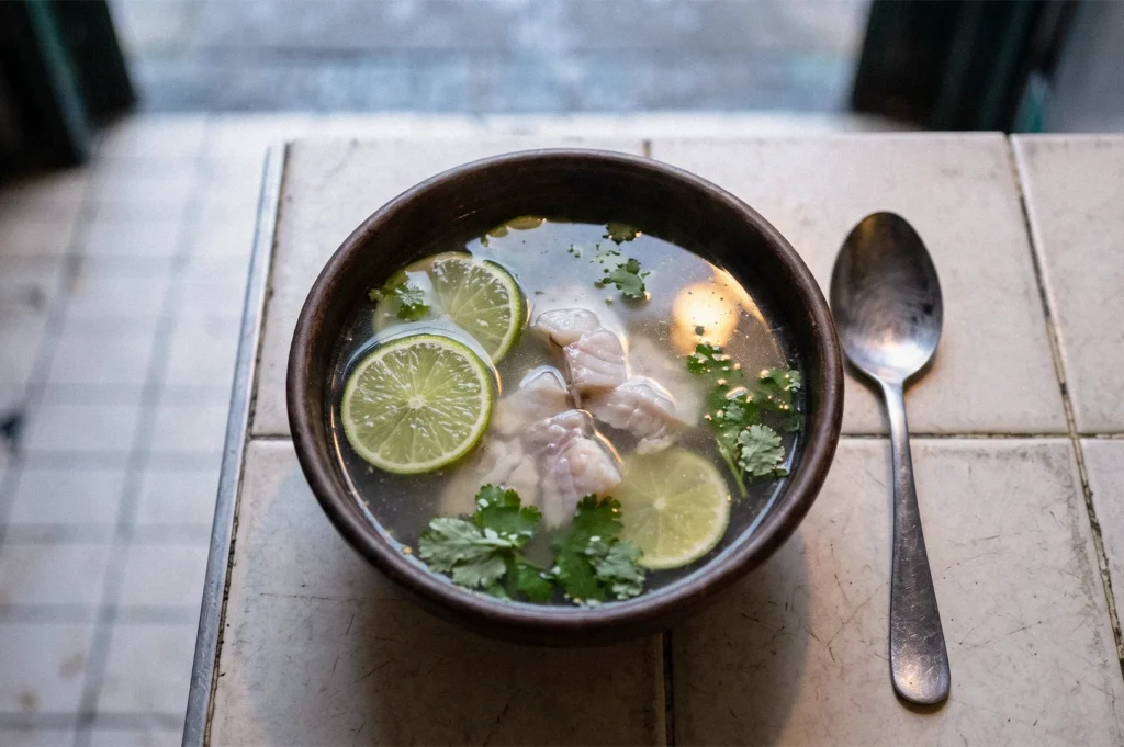 A bowl of clear soup with sliced fish, lime rounds, cilantro, and herbs, served with a spoon on a tiled table.