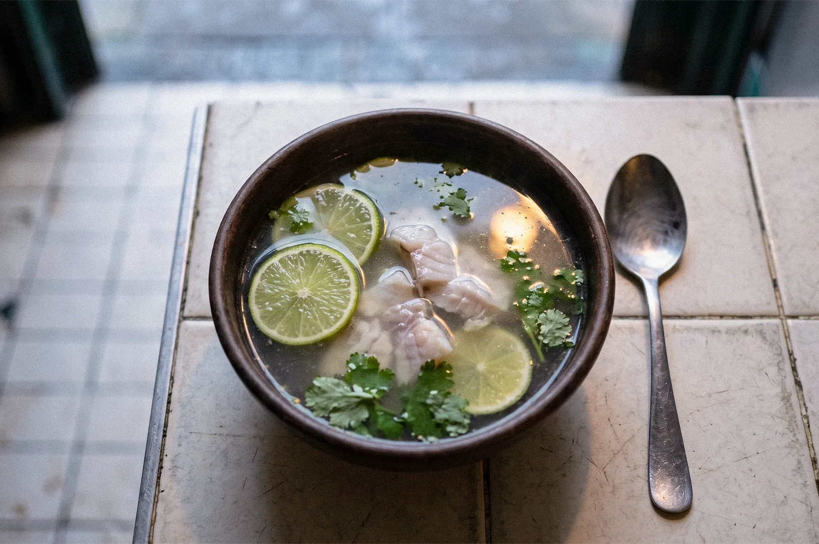 A bowl of clear soup with sliced fish, lime rounds, cilantro, and herbs, served with a spoon on a tiled table.