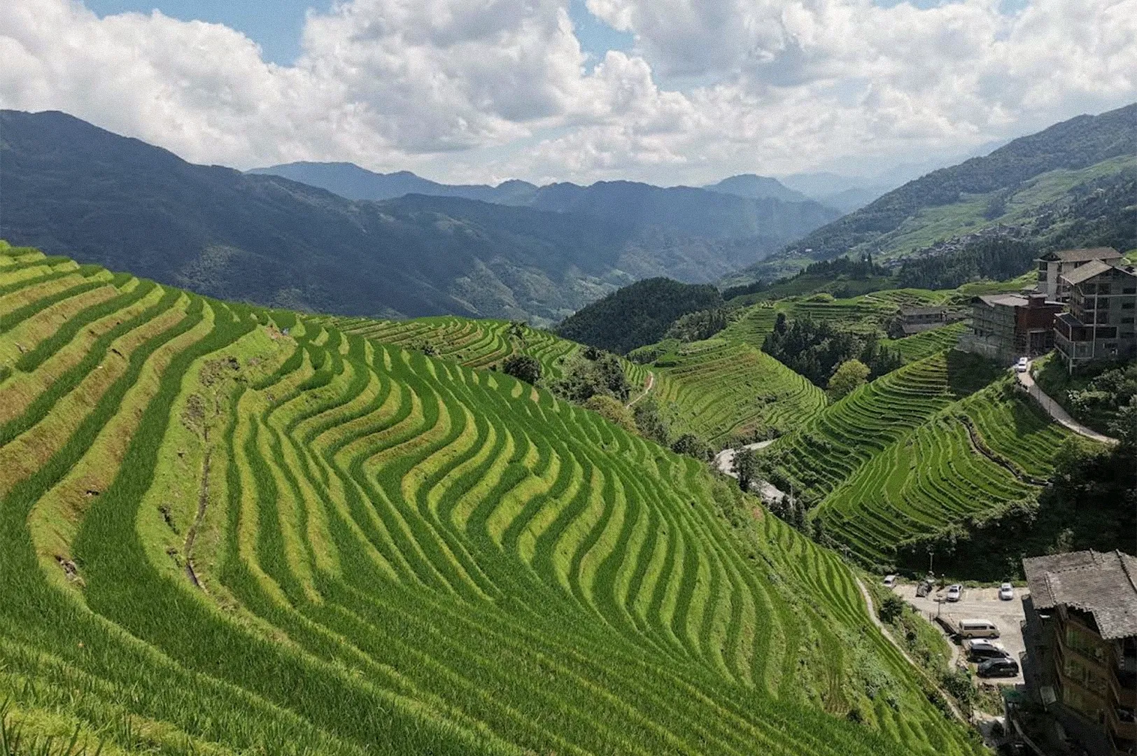 A sweeping view of lush green rice terraces curving across mountain slopes, with traditional buildings along the hillside, layered mountain ranges in the distance, and fluffy white clouds overhead.