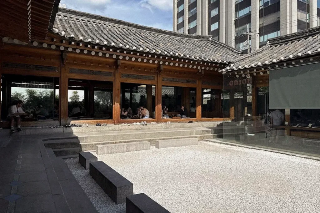 A traditional Korean-style café features dark wooden window frames, curved gray tiled roofs, stone steps, and a white gravel courtyard, with modern high-rise buildings visible behind it.