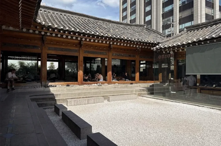 A traditional Korean-style café features dark wooden window frames, curved gray tiled roofs, stone steps, and a white gravel courtyard, with modern high-rise buildings visible behind it.