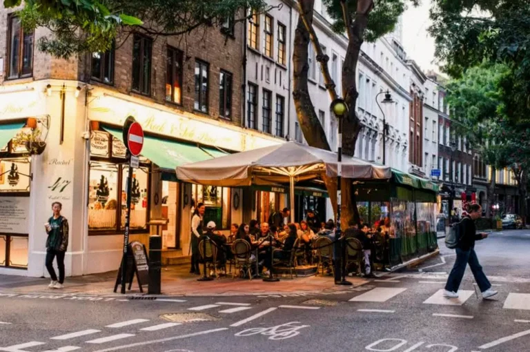 A bustling London street with a corner café offering outdoor dining under beige umbrellas, where diners sit along the sidewalk as pedestrians cross the road.