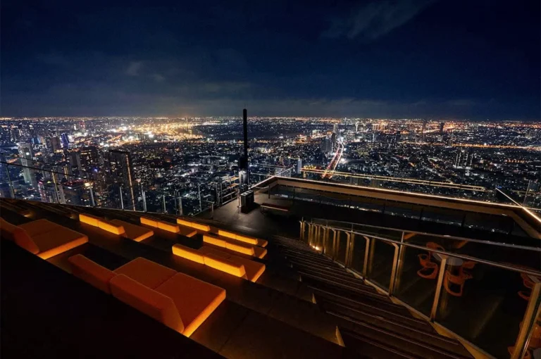Nighttime view of Bangkok’s illuminated skyline from a rooftop terrace, with city lights stretching into the distance and warmly lit seating in the foreground.