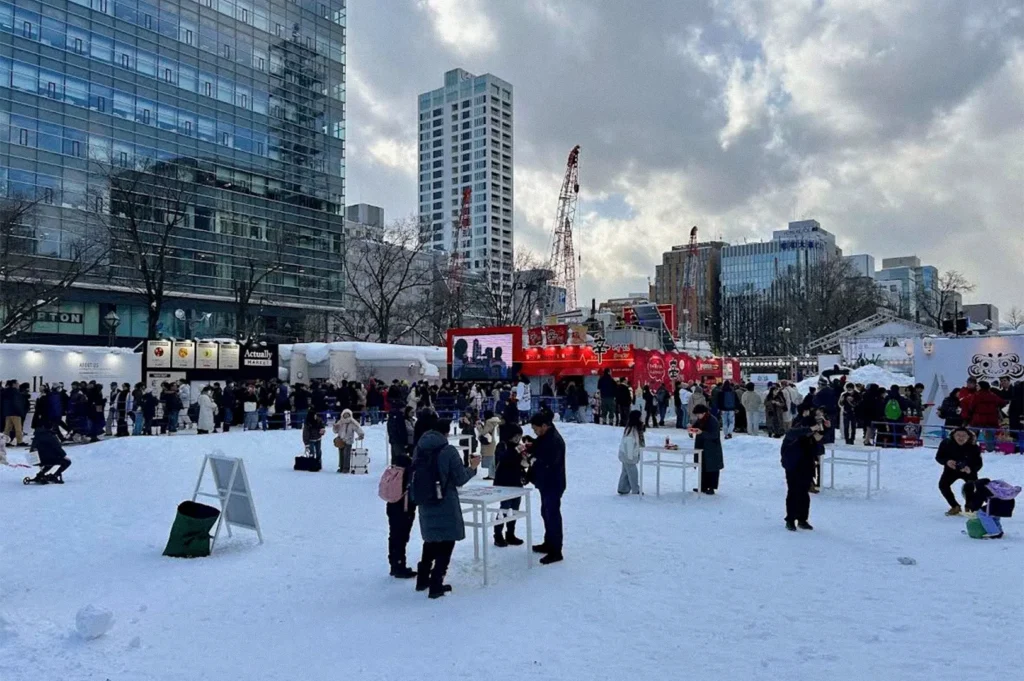 Visitors explore a snow-covered winter festival in an urban plaza in Hokkaido, with snow displays, food stalls, a large digital screen, and modern buildings surrounding the event.