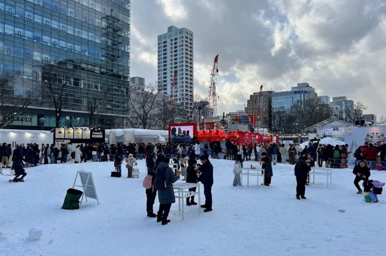 Visitors explore a snow-covered winter festival in an urban plaza in Hokkaido, with snow displays, food stalls, a large digital screen, and modern buildings surrounding the event.
