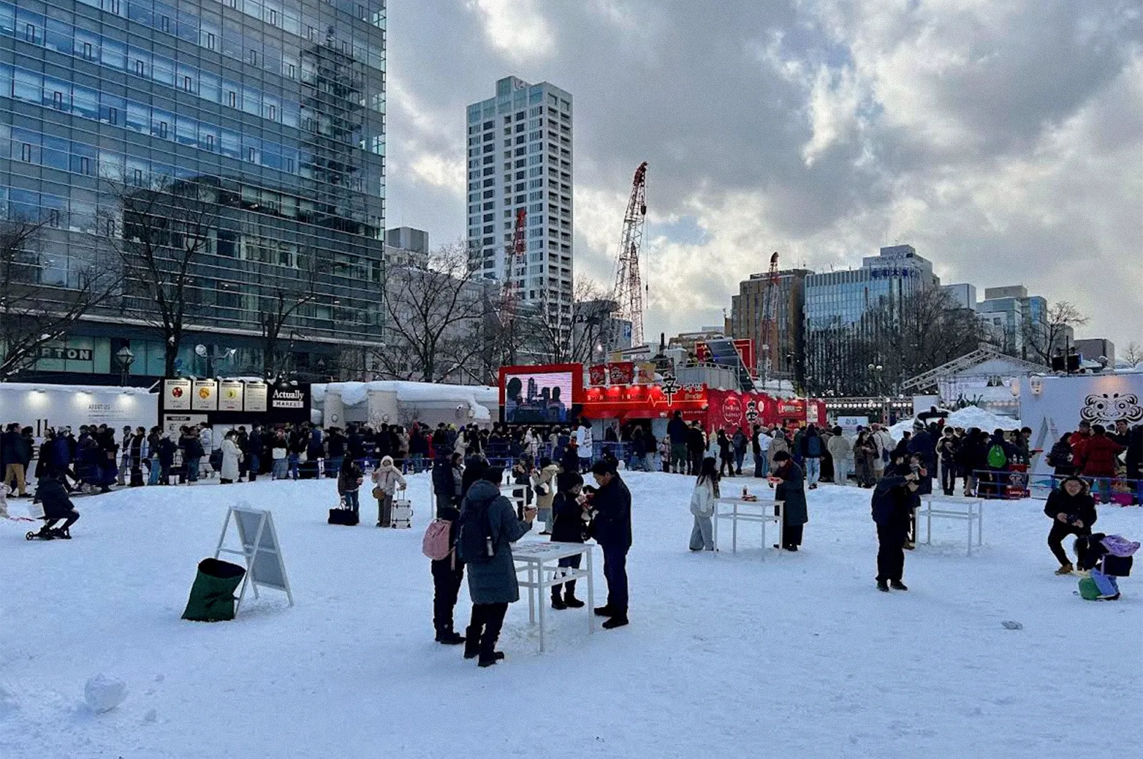 Visitors explore a snow-covered winter festival in an urban plaza in Hokkaido, with snow displays, food stalls, a large digital screen, and modern buildings surrounding the event.