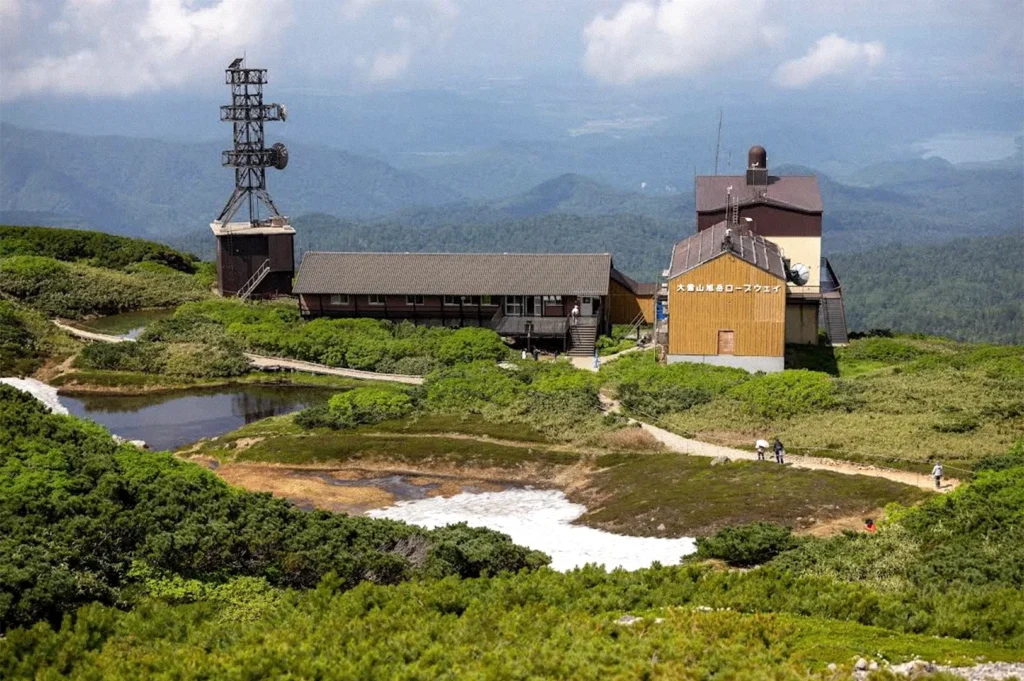 A mountain ropeway station surrounded by green alpine vegetation, a small pond, and winding walking trails, with layers of forested mountains stretching into the distance beneath a partly cloudy sky.