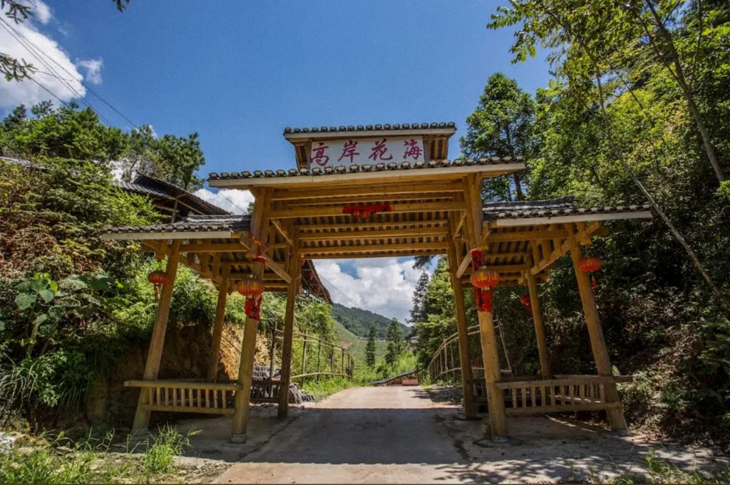 A traditional Chinese wooden gateway adorned with red lanterns and Chinese characters frames a paved pathway leading into a green mountain valley beneath a bright blue sky.