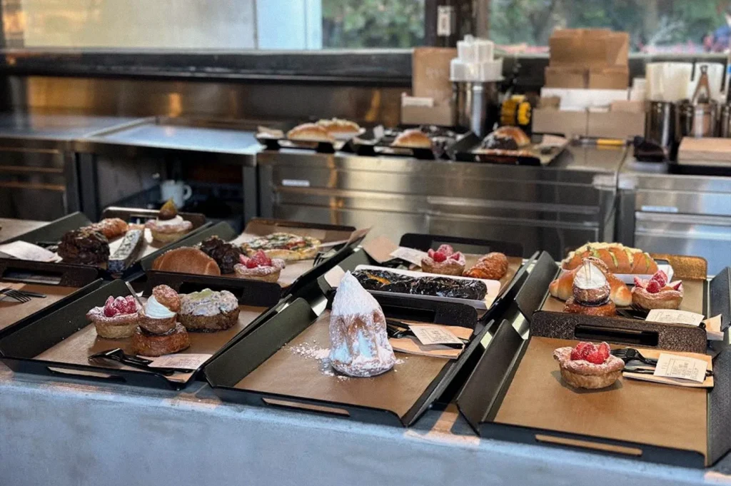 An array of pastries, including fruit tarts, cream-filled desserts, chocolate pastries, and freshly baked breads, is arranged on black trays along a bakery counter.