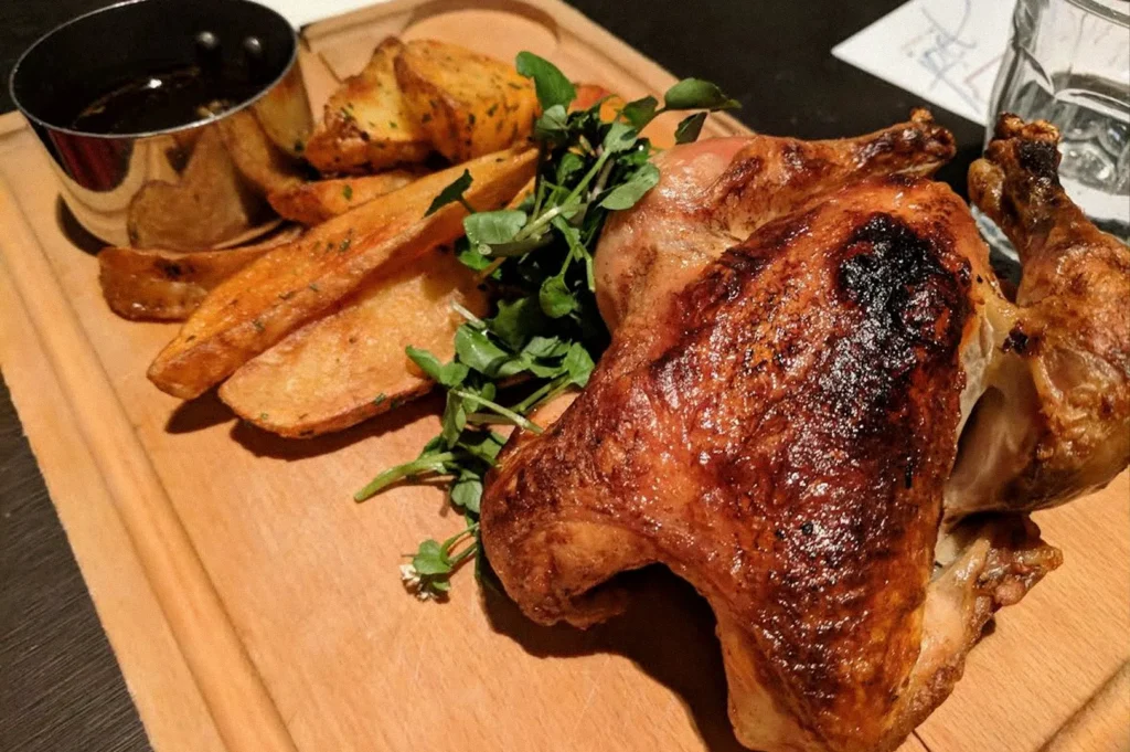 Golden-brown roasted chicken served on a wooden board with seasoned potato wedges and fresh greens.