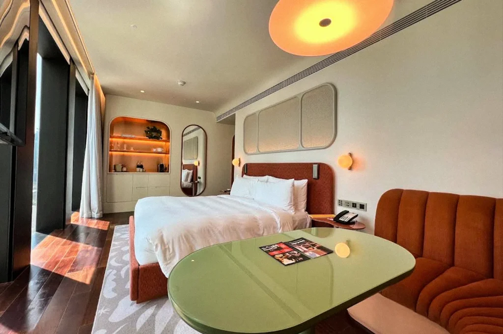 Modern hotel room with a white bed, orange upholstered headboard and armchair, green table, and warm ambient lighting beside floor-to-ceiling windows.