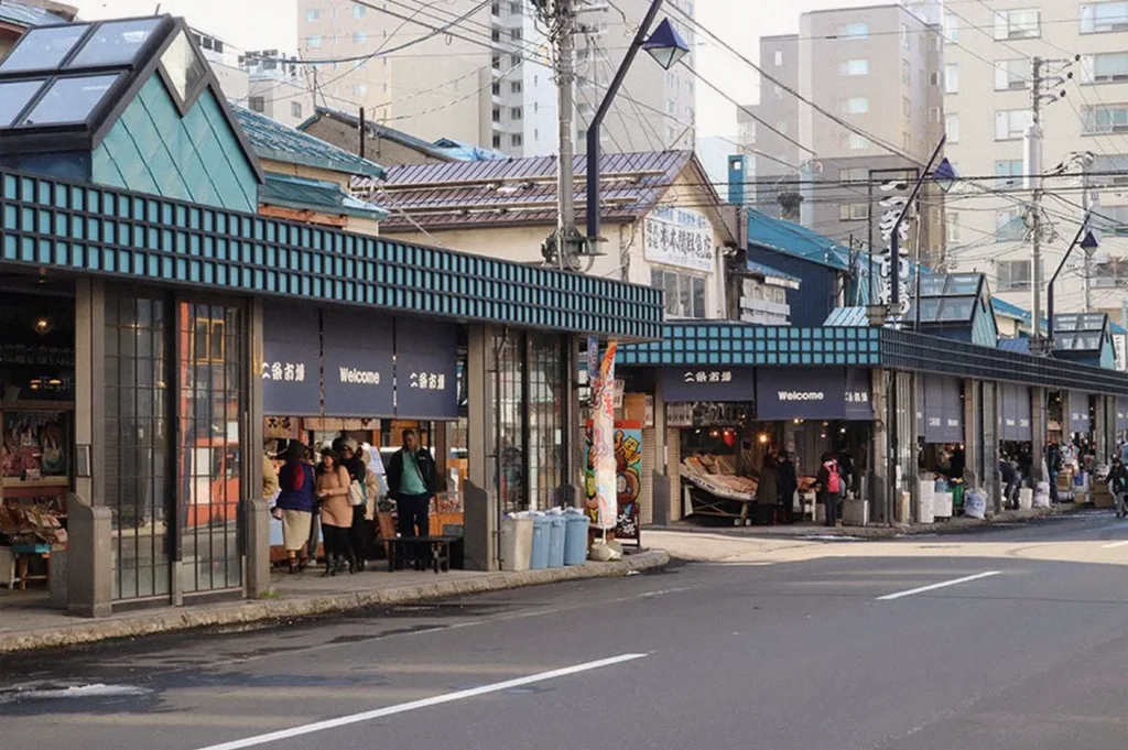 A row of traditional Japanese market stalls with blue and teal roof accents, shop signs, and open storefronts selling fresh seafood and local goods along a city street in Hokkaido.