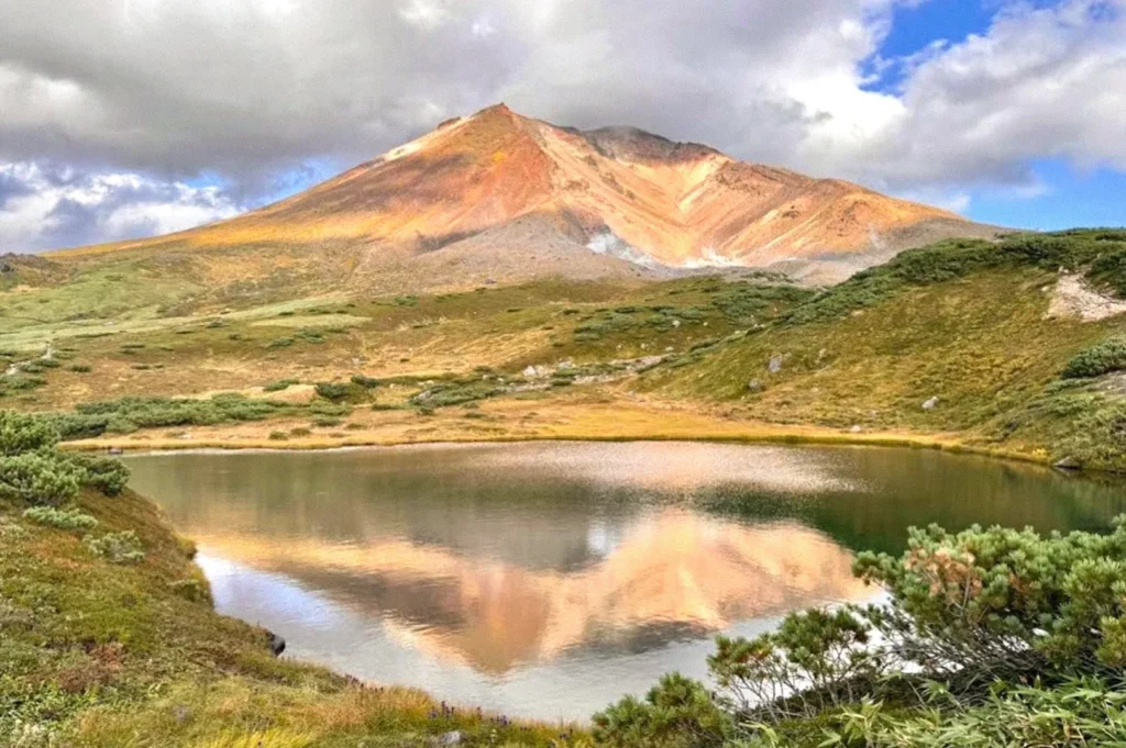 A sunlit mountain with golden and reddish slopes reflected in a calm alpine lake, surrounded by green tundra beneath dramatic clouds