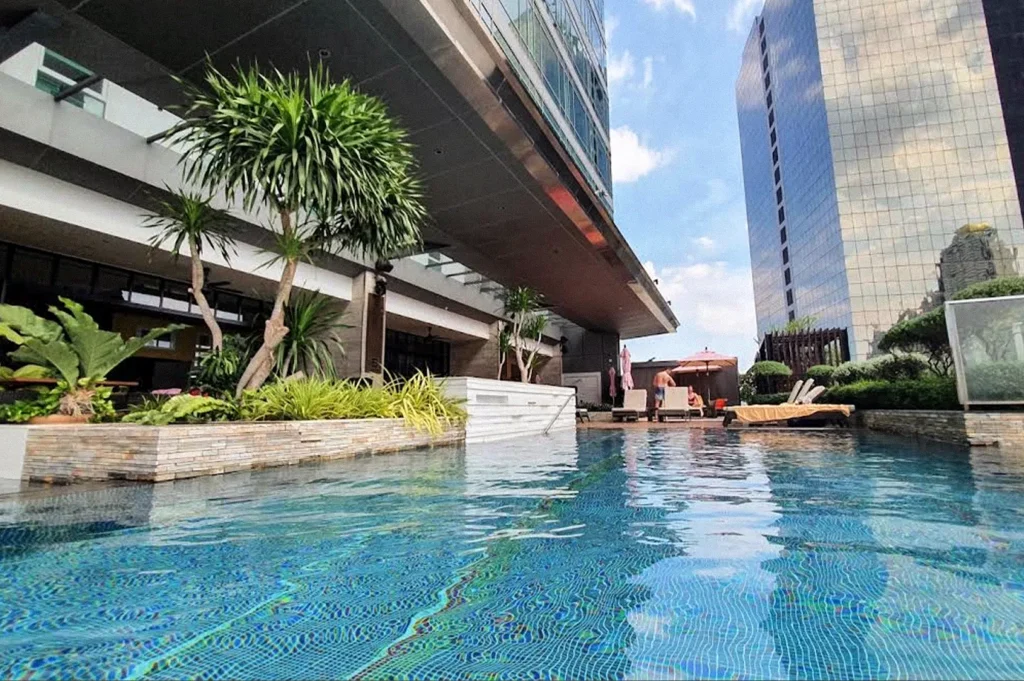 Rooftop swimming pool at Eastin Grand Hotel Sathorn, surrounded by tropical plants, lounge chairs, and modern high-rise buildings.