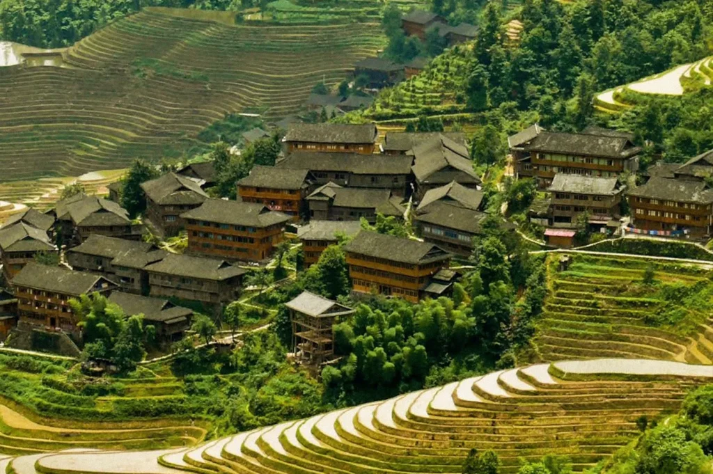 A cluster of traditional wooden houses with dark sloping roofs sits among lush greenery and expansive rice terraces carved into the surrounding hills.