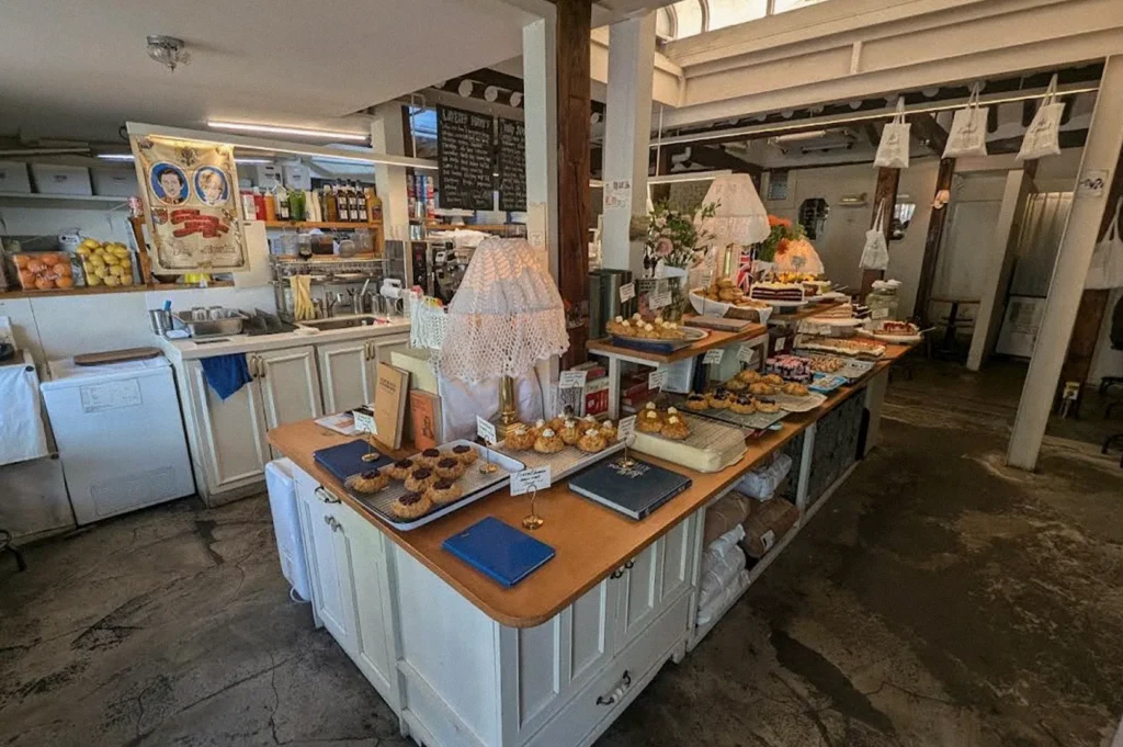 A cozy bakery café displays assorted cakes, cookies, and pastries on wooden tables, with decorative lamps, handwritten menu boards, and a service counter in the background.