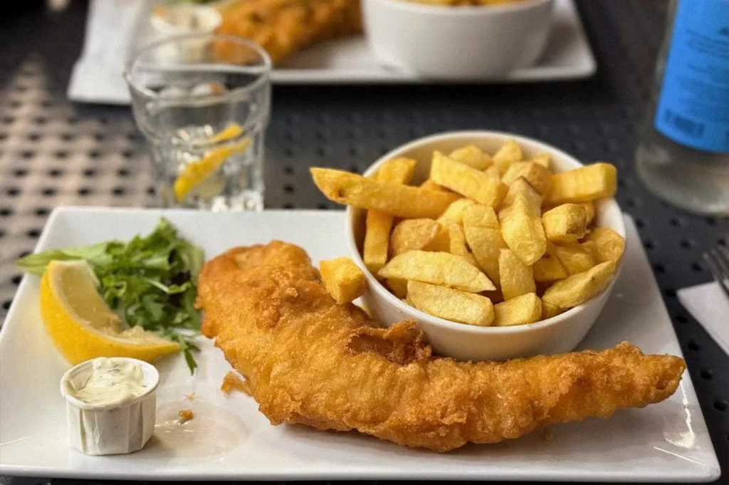 Classic fish and chips featuring a large piece of crispy battered fish, thick-cut fries, lemon wedges, fresh greens, and tartar sauce on a white plate.