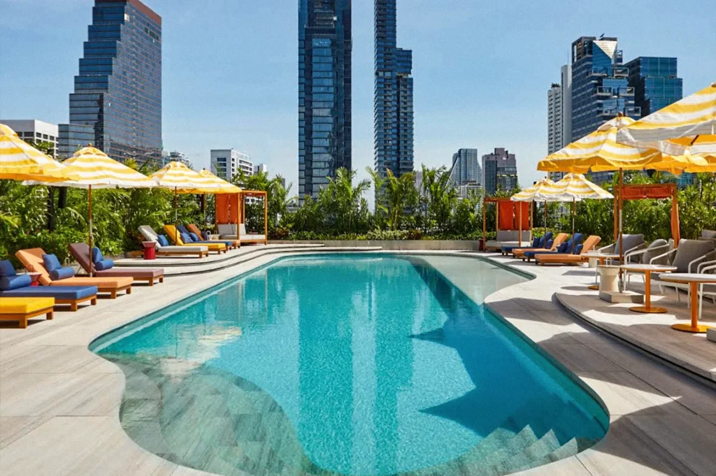 Rooftop swimming pool surrounded by yellow-striped umbrellas, colorful lounge chairs, lush greenery, and Bangkok skyscrapers under a clear blue sky.