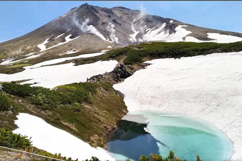A turquoise alpine pond bordered by extensive patches of lingering snow, with a rugged volcanic mountain emitting wisps of steam in the background.